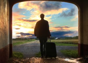 Silhouette of man pulling wheeled luggage, walking toward the sunset. Orange and pink clouds fill a vista that is full of possibilities.